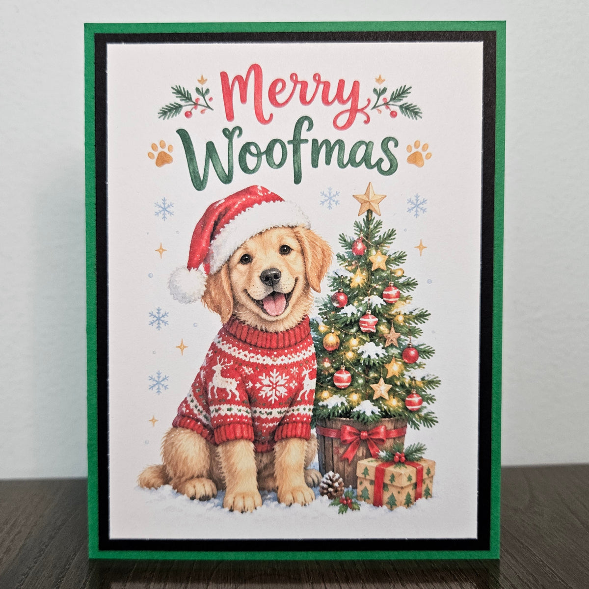 A Christmas greeting card featuring a golden retriever puppy wearing a red Santa hat and red knit sweater, sitting beside a decorated Christmas tree and wrapped presents. The card reads “Merry Woofmas” in red and green lettering, with paw prints and holly accents.