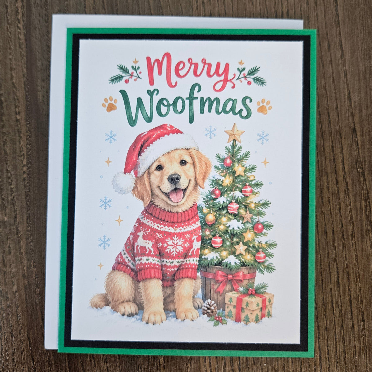 A Christmas greeting card featuring a golden retriever dog wearing a red Santa hat and red knit sweater, sitting beside a decorated Christmas tree. The card reads 'Merry Woofmas' in red and green lettering, with paw prints and snowflake accents.