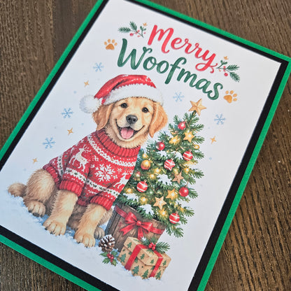 A Christmas greeting card featuring an illustration of a golden retriever wearing a red Santa hat and red holiday sweater, sitting beside a decorated Christmas tree and wrapped presents. The card reads “Merry Woofmas” in red and green lettering.