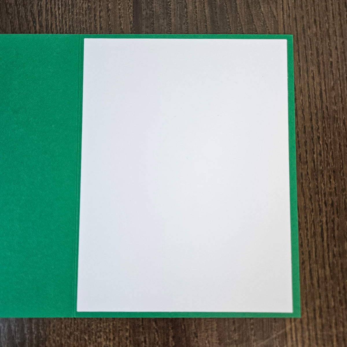 A white card or sheet of paper placed on top of a green card or folder, photographed from above on a dark wood surface.