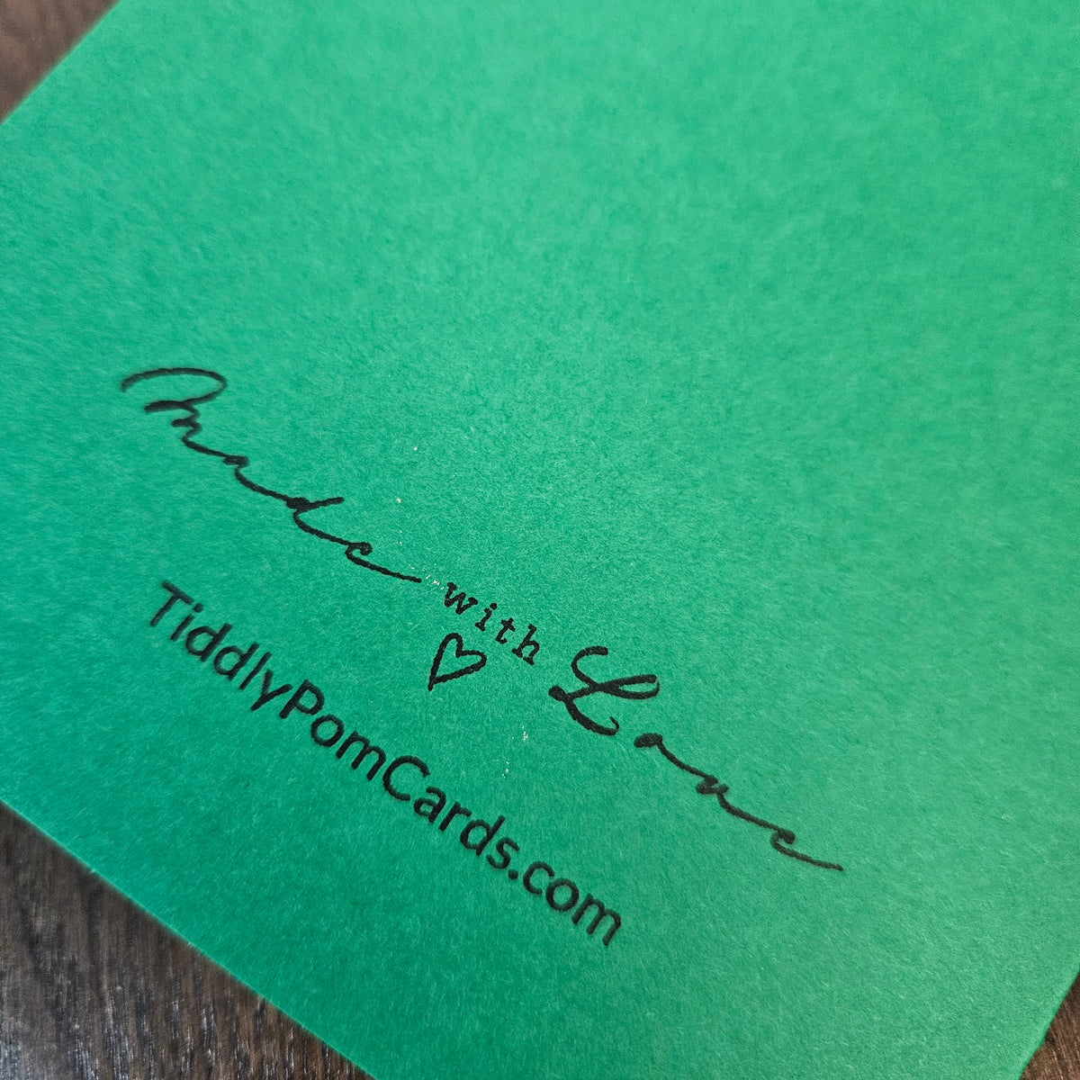 A close-up of a bright green textured card with black printed text reading 'Made with Love' and 'TiddlyPomCards.com' on a dark wood surface.