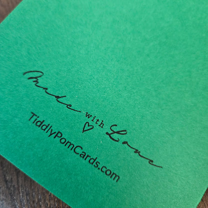 A close-up of a bright green textured card with black printed text reading 'Made with Love' and 'TiddlyPomCards.com' on a dark wood surface.