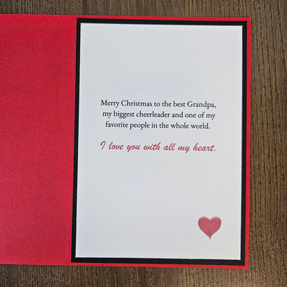 An open red greeting card with a white front insert printed with a Christmas message for a grandpa and a small pink heart, photographed on a wood surface.