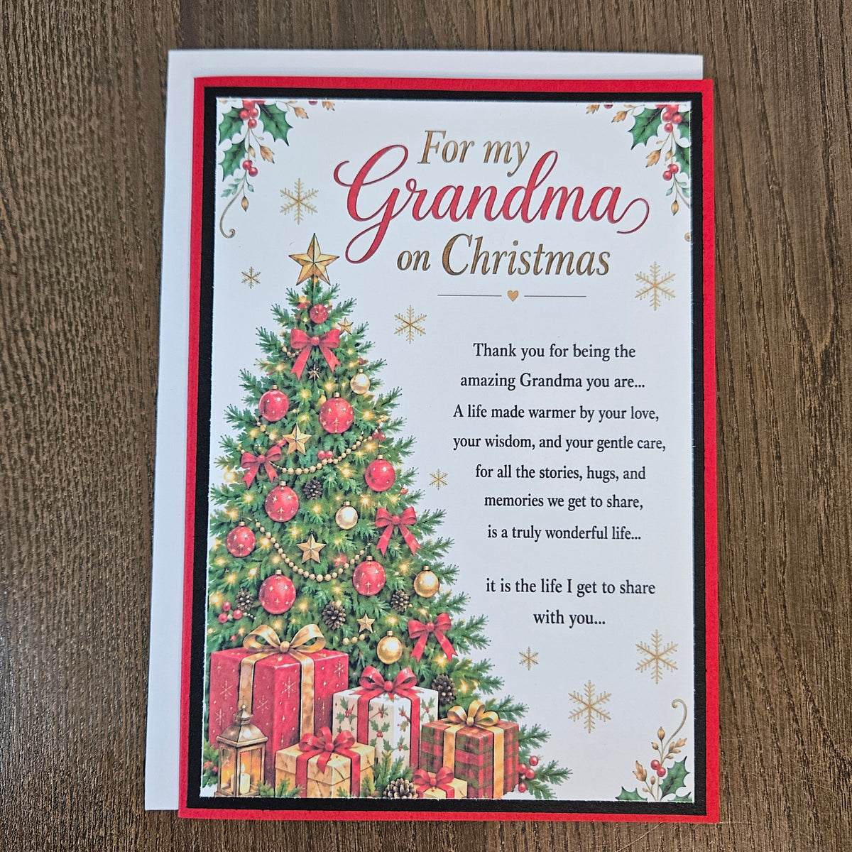 A Christmas-themed greeting card addressed 'For my Grandma on Christmas' lies flat on a dark wood surface. The card features a decorated Christmas tree, wrapped gifts, and a red border, with a heartfelt thank you message printed on a white background.