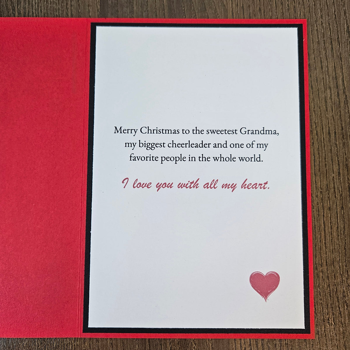 A Christmas greeting card with a red textured cover and a white inner card featuring a message for 'the sweetest Grandma' in black and red text, with a small pink heart on a wood background.