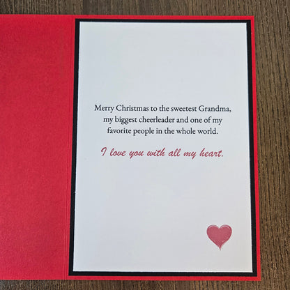 A Christmas greeting card with a red textured cover and a white inner card featuring a message for 'the sweetest Grandma' in black and red text, with a small pink heart on a wood background.