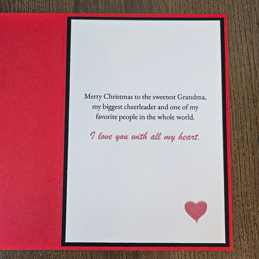 A Christmas greeting card with a red textured cover and a white inner card featuring a message for 'the sweetest Grandma' in black and red text, with a small pink heart on a wood background.