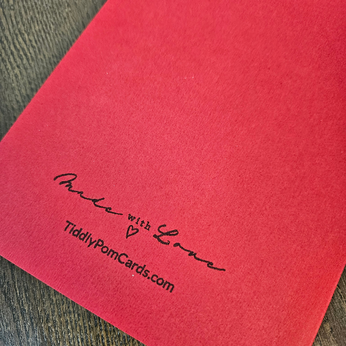 A close-up of a red textured card or envelope corner printed with the words 'Made with Love' and the website 'TiddlyPomCards.com' in black script, placed on a dark wood surface.