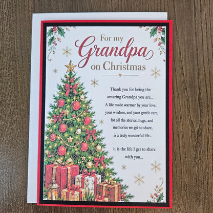 A Christmas-themed card for a grandpa, featuring a decorated Christmas tree, wrapped gifts, and festive text on a white card with a red border, placed on a wooden surface.