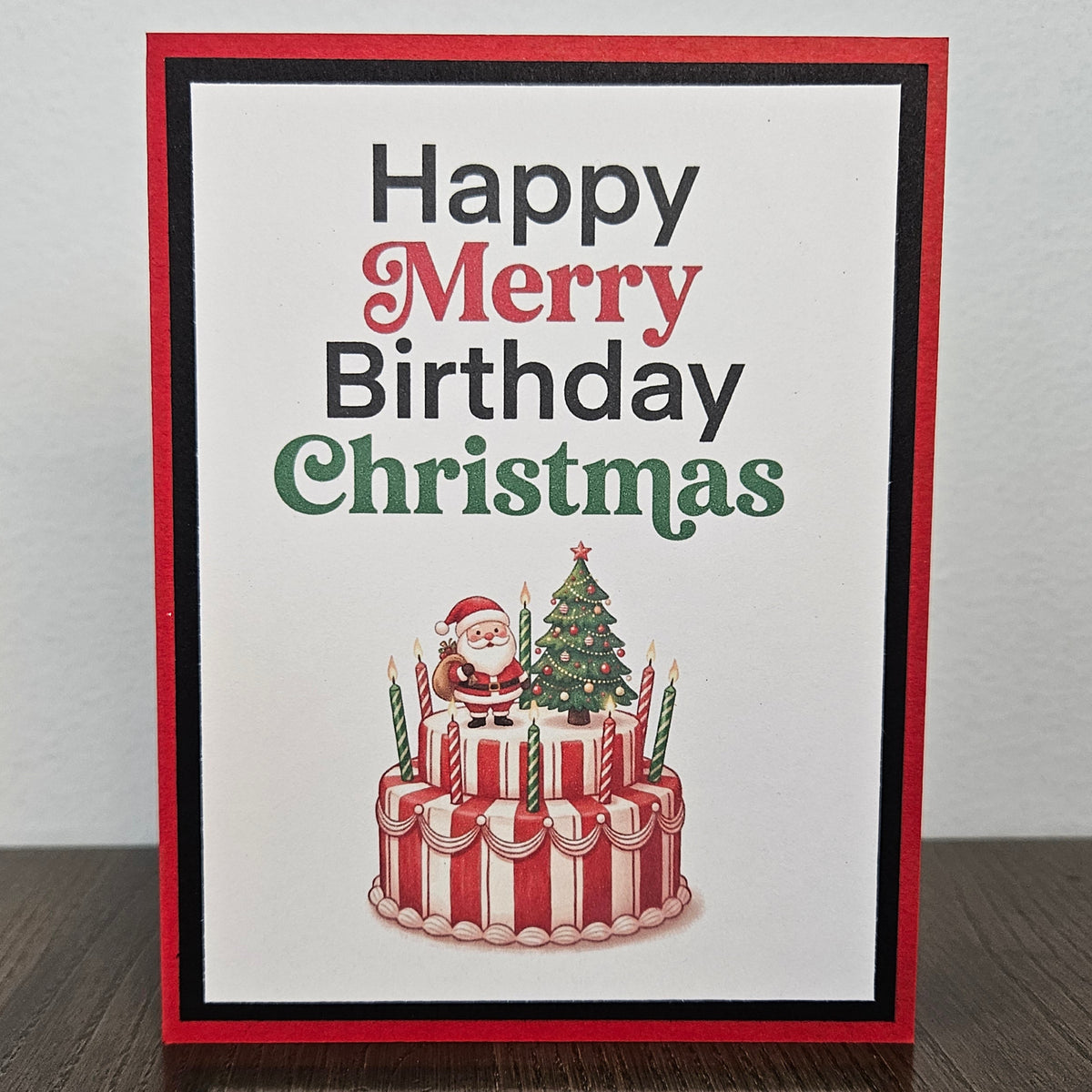 A handmade greeting card with a red border and a white center, reading 'Happy Merry Birthday Christmas' in black, red, and green lettering above an illustration of a red-and-white striped birthday cake with candles, Santa Claus, and a Christmas tree. The card is photographed standing upright on a dark wooden surface against a light gray wall.