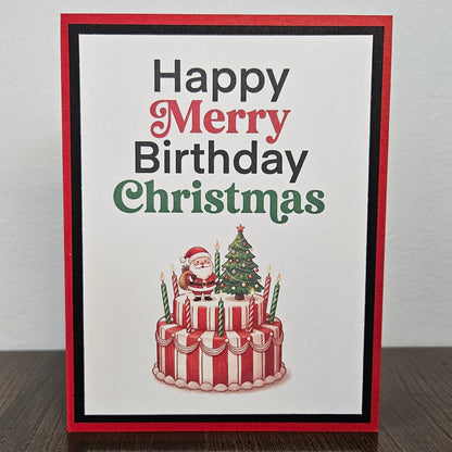 A handmade greeting card with a red border and a white center, reading 'Happy Merry Birthday Christmas' in black, red, and green lettering above an illustration of a red-and-white striped birthday cake with candles, Santa Claus, and a Christmas tree. The card is photographed standing upright on a dark wooden surface against a light gray wall.