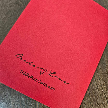 A bright red textured card or envelope cover printed with the words 'Made with Love' and 'TiddlyPomCards.com' in black script, photographed on a dark wood surface.