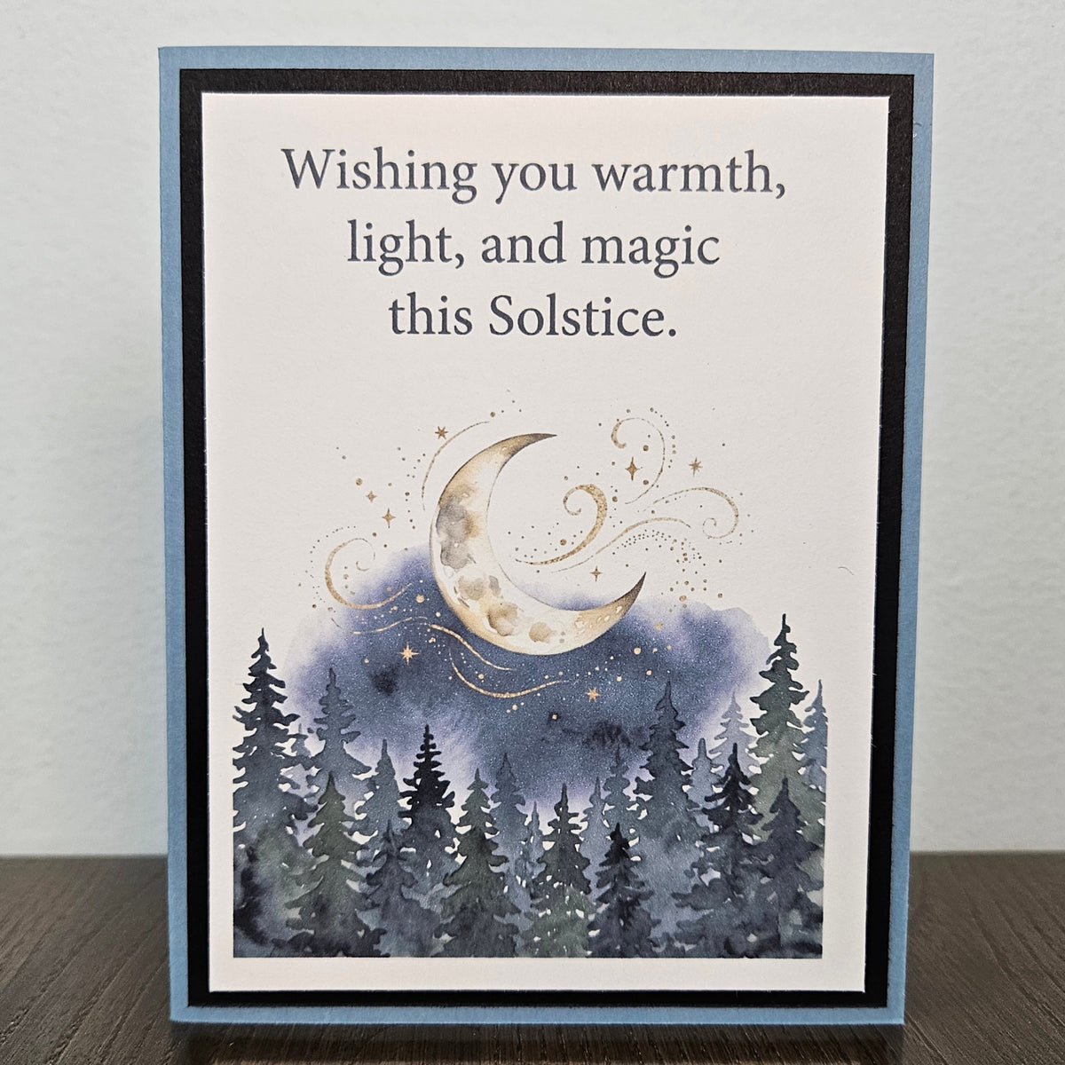 A handmade greeting card with a blue border, featuring the text 'Wishing you warmth, light, and magic this Solstice.' above an illustration of a crescent moon, stars, and a dark blue forest.