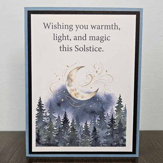 A handmade greeting card with a blue border, featuring the text 'Wishing you warmth, light, and magic this Solstice.' above an illustration of a crescent moon, stars, and a dark blue forest.