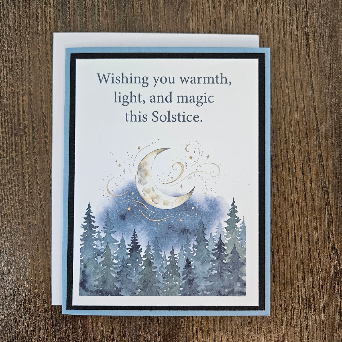Full front of the handmade Winter Solstice card with a white envelope behind it, showing the moonlit evergreen forest design and printed sentiment.