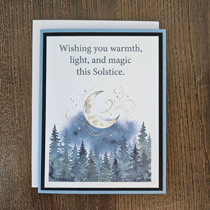 Full front of the handmade Winter Solstice card with a white envelope behind it, showing the moonlit evergreen forest design and printed sentiment.