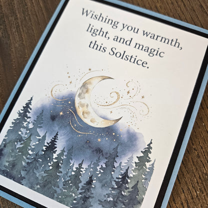 Angled close-up of the Winter Solstice card showing the crescent moon, stars, evergreen forest, and printed greeting.