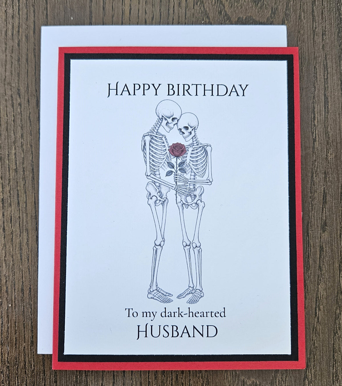 Red and black skeleton-couple birthday card for a husband shown with its included white envelope.