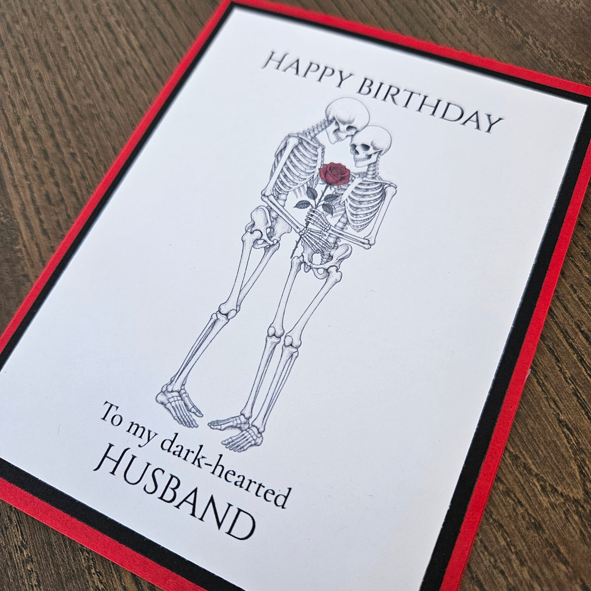 Angled close-up of the embracing skeleton couple, red rose, husband birthday sentiment, and layered red and black borders.