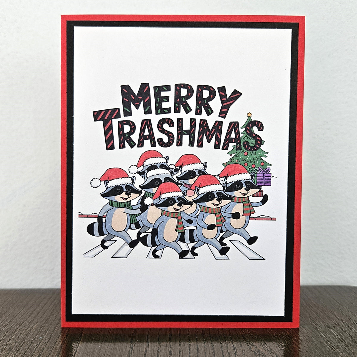 A handmade greeting card featuring a cartoon group of raccoons wearing Santa hats and scarves, walking across a crosswalk with a Christmas tree and presents. The text 'MERRY TRASHMAS' is written above them.