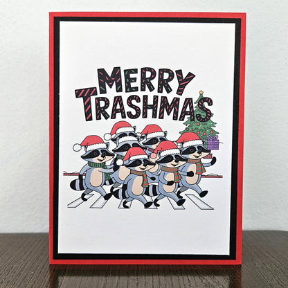 A handmade greeting card featuring a cartoon group of raccoons wearing Santa hats and scarves, walking across a crosswalk with a Christmas tree and presents. The text 'MERRY TRASHMAS' is written above them.