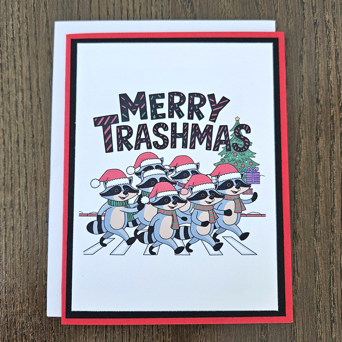 A festive greeting card featuring the text 'MERRY TRASHMAS' above a group of cartoon raccoons wearing Santa hats and sunglasses, walking across a zebra crossing. The card has a red border, a black inner border, and is shown with a white backing card on a dark wood background.