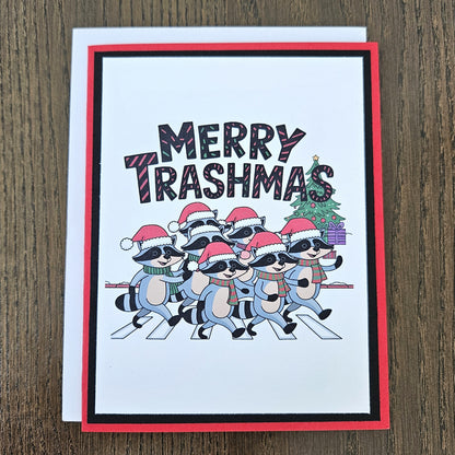 A festive greeting card featuring the text 'MERRY TRASHMAS' above a group of cartoon raccoons wearing Santa hats and sunglasses, walking across a zebra crossing. The card has a red border, a black inner border, and is shown with a white backing card on a dark wood background.