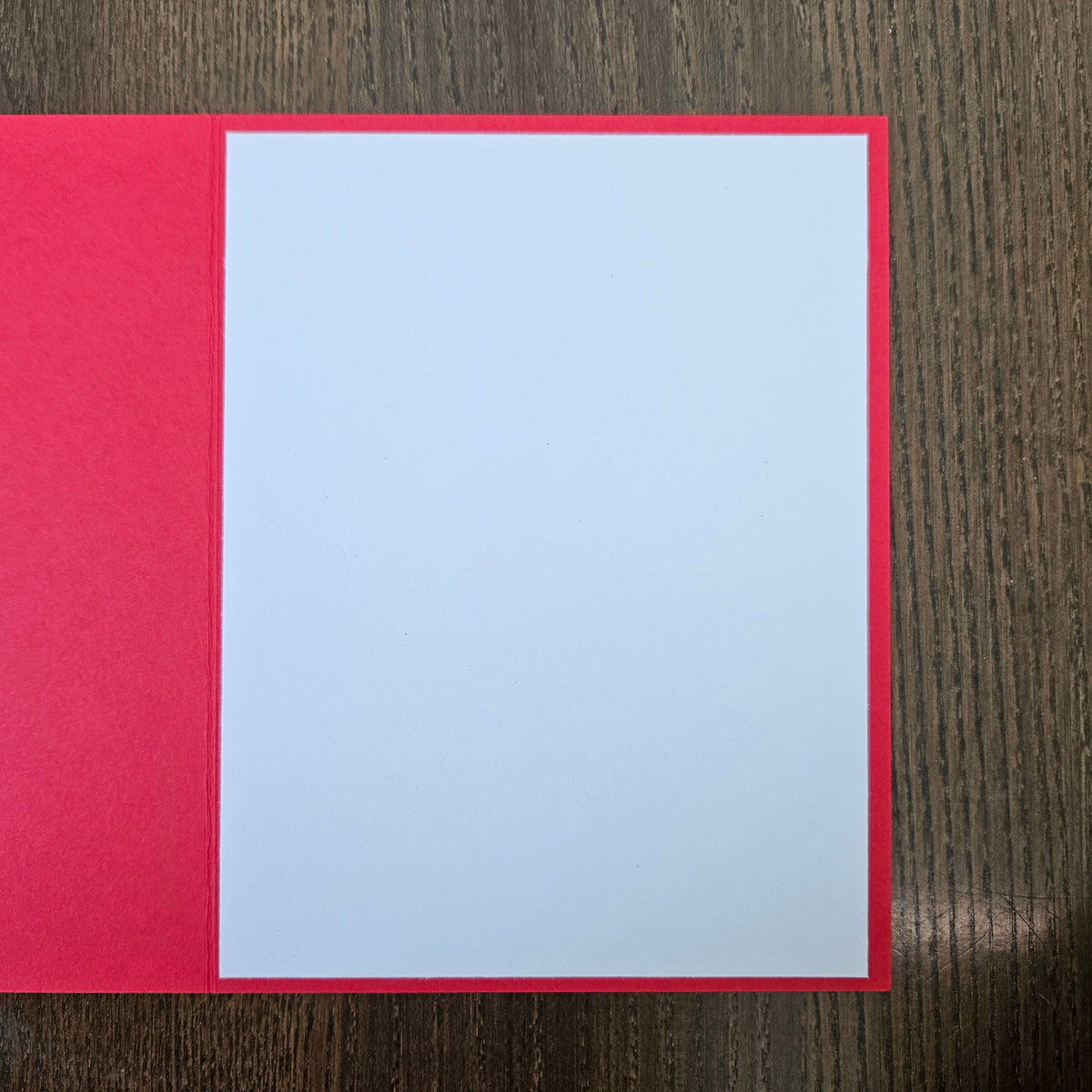 A blank white rectangular card or paper sheet with a bright red border, placed on a dark brown wood-grain surface.
