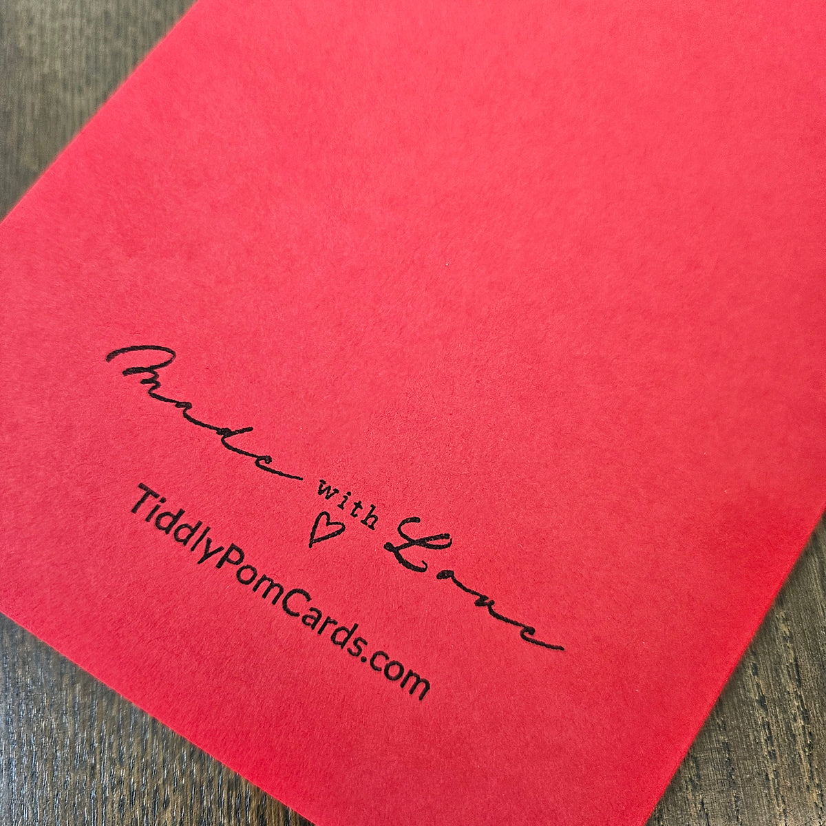 A close-up of a bright red square card or envelope with black printed text reading 'Made with Love' and 'TiddlyPomCards.com' on a dark wood surface.