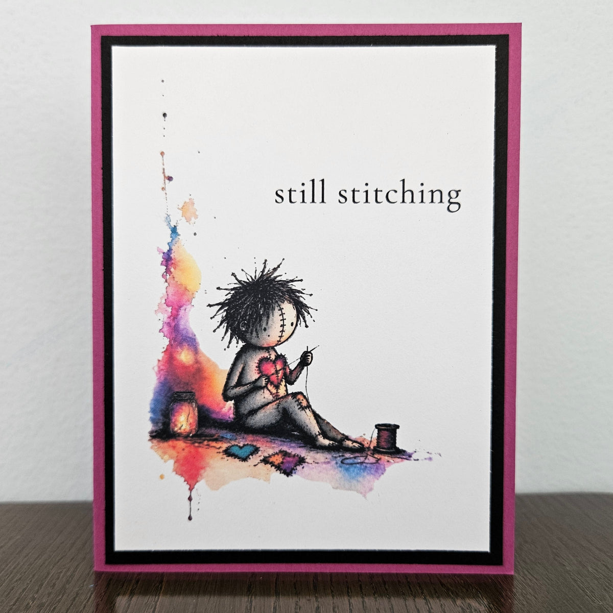 Handmade magenta and black encouragement card with still stitching wording, a stitched doll mending a pink heart, and colorful printed splashes.