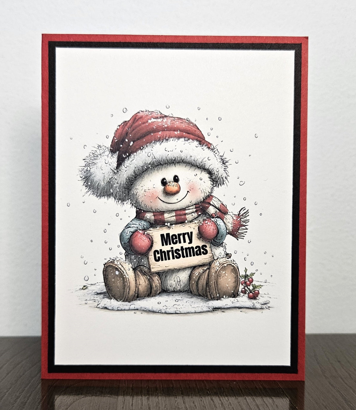 Handmade red and black Christmas card with a smiling snowman in a Santa hat and scarf holding a Merry Christmas sign.