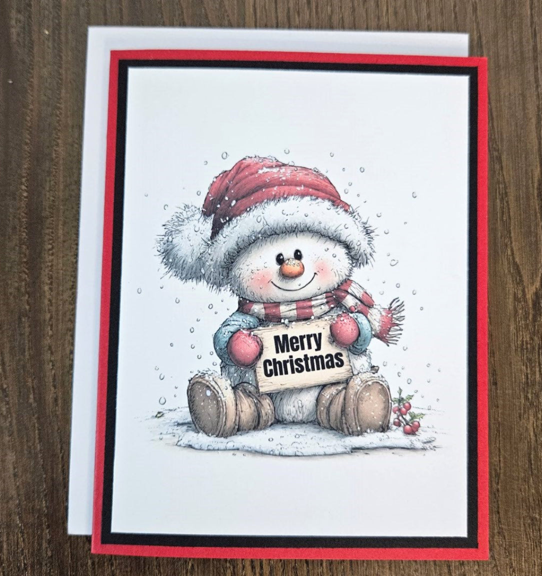 Red and black snowman Christmas card shown with its included white envelope on a wooden surface.