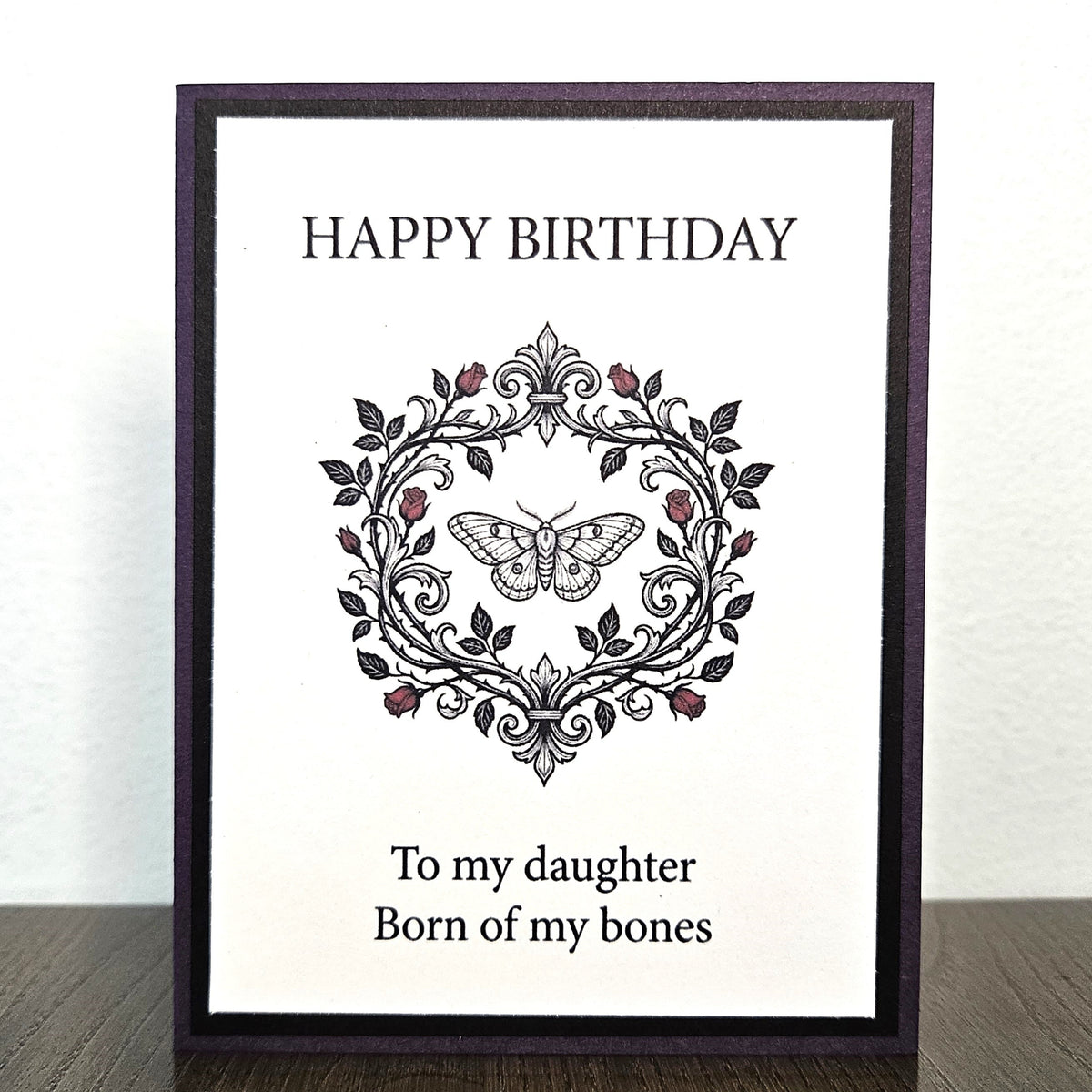 A framed birthday card standing on a dark wooden surface against a white wall. The card has a dark purple frame and a white background with the text 'HAPPY BIRTHDAY' at the top and 'To my daughter Born of my bones' at the bottom, centered around an ornate black floral wreath with a butterfly and small red rose accents.