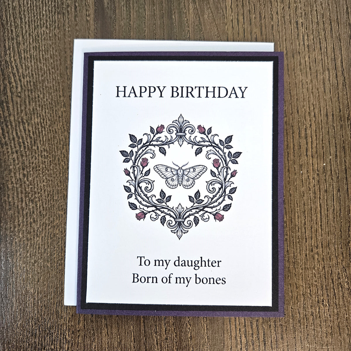 A handmade birthday card with a white front and a dark purple border, placed on a wooden surface. The card reads “HAPPY BIRTHDAY” above an ornate floral and butterfly illustration, and below the illustration it says “To my daughter Born of my bones.