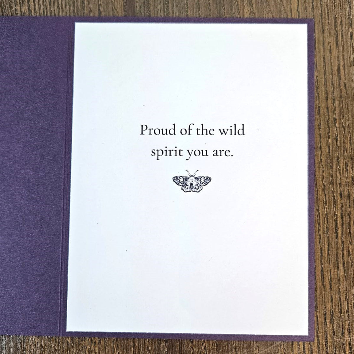 A greeting card with a white center panel and textured purple border, placed on a wooden surface. The card reads, 'Proud of the wild spirit you are.