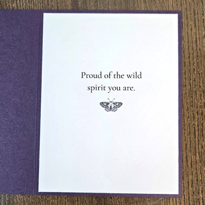 A greeting card with a white center panel and textured purple border, placed on a wooden surface. The card reads, 'Proud of the wild spirit you are.