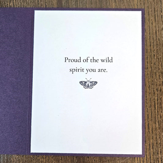 A greeting card with a white center panel and textured purple border, placed on a wooden surface. The card reads, 'Proud of the wild spirit you are.