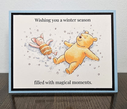 Handmade landscape pale blue and black Winnie the Pooh card showing Pooh and Piglet making snow angels, with winter season and magical moments wording.