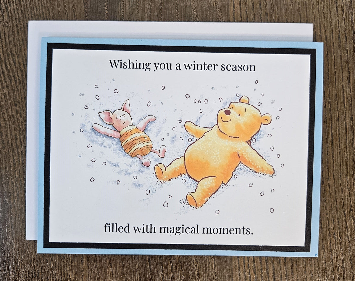 Landscape Pooh and Piglet snow-angel winter card laid on a wooden surface with its included white envelope.