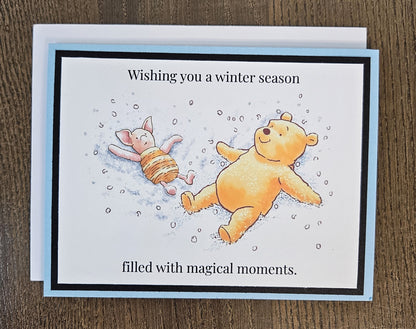 Landscape Pooh and Piglet snow-angel winter card laid on a wooden surface with its included white envelope.