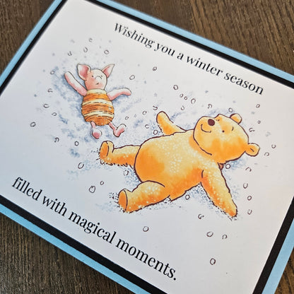 Angled close-up of Pooh and Piglet making snow angels, the printed winter greeting, and pale blue and black layered cardstock borders.
