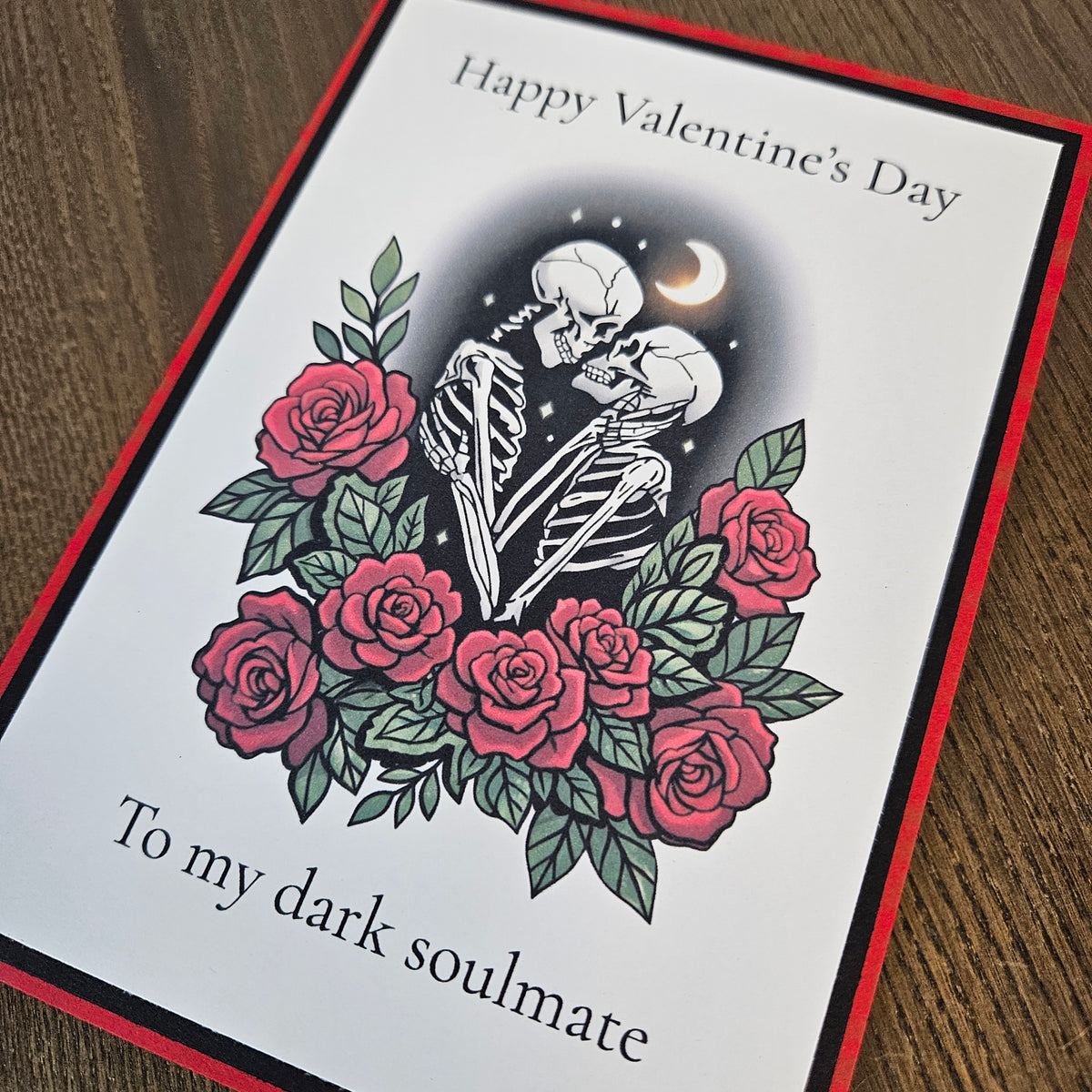 Angled detail of the skeleton couple, pink-red roses, green foliage and crescent moon on the layered gothic Valentine card.