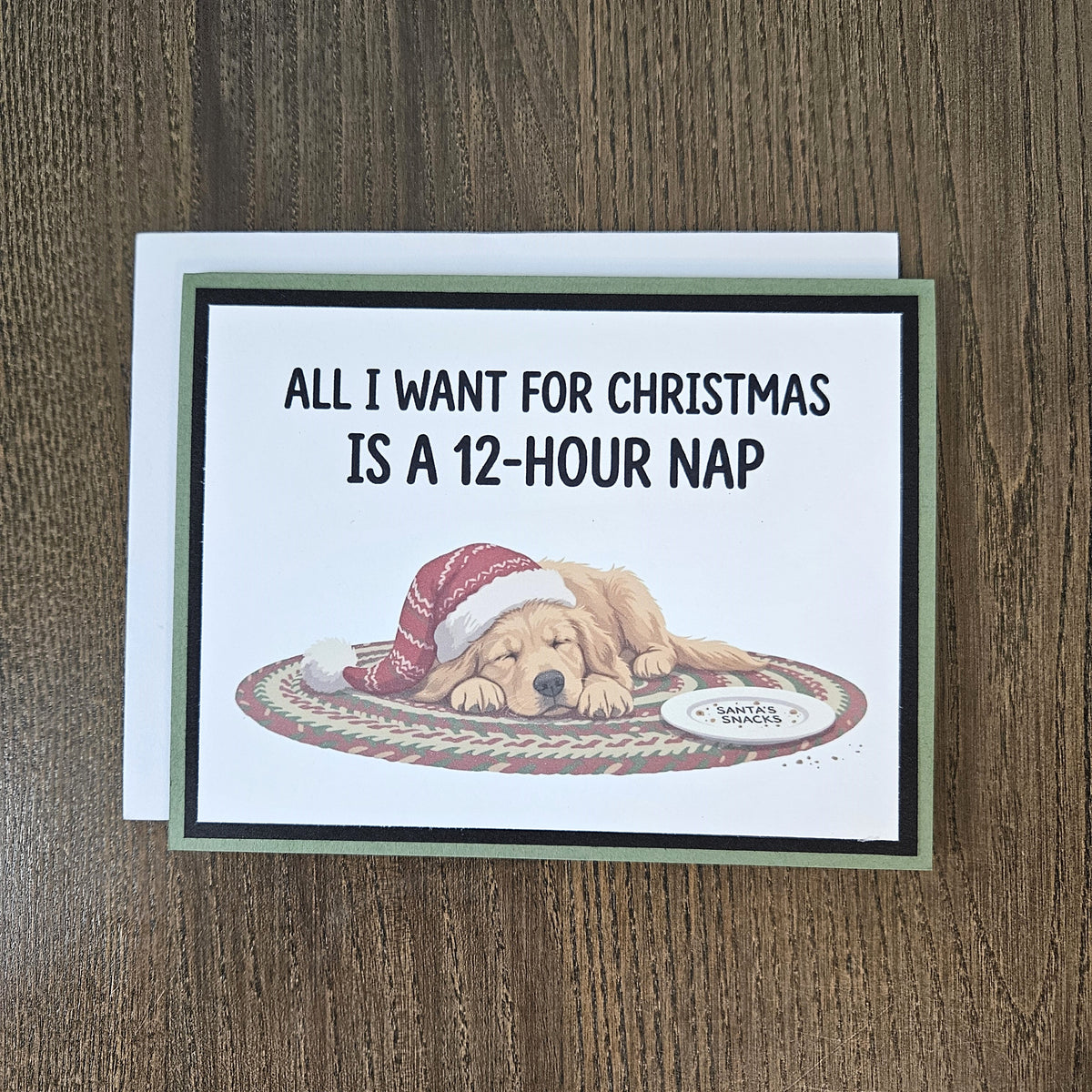 Green and black sleeping-dog Christmas card shown with its included white envelope on a wooden surface.