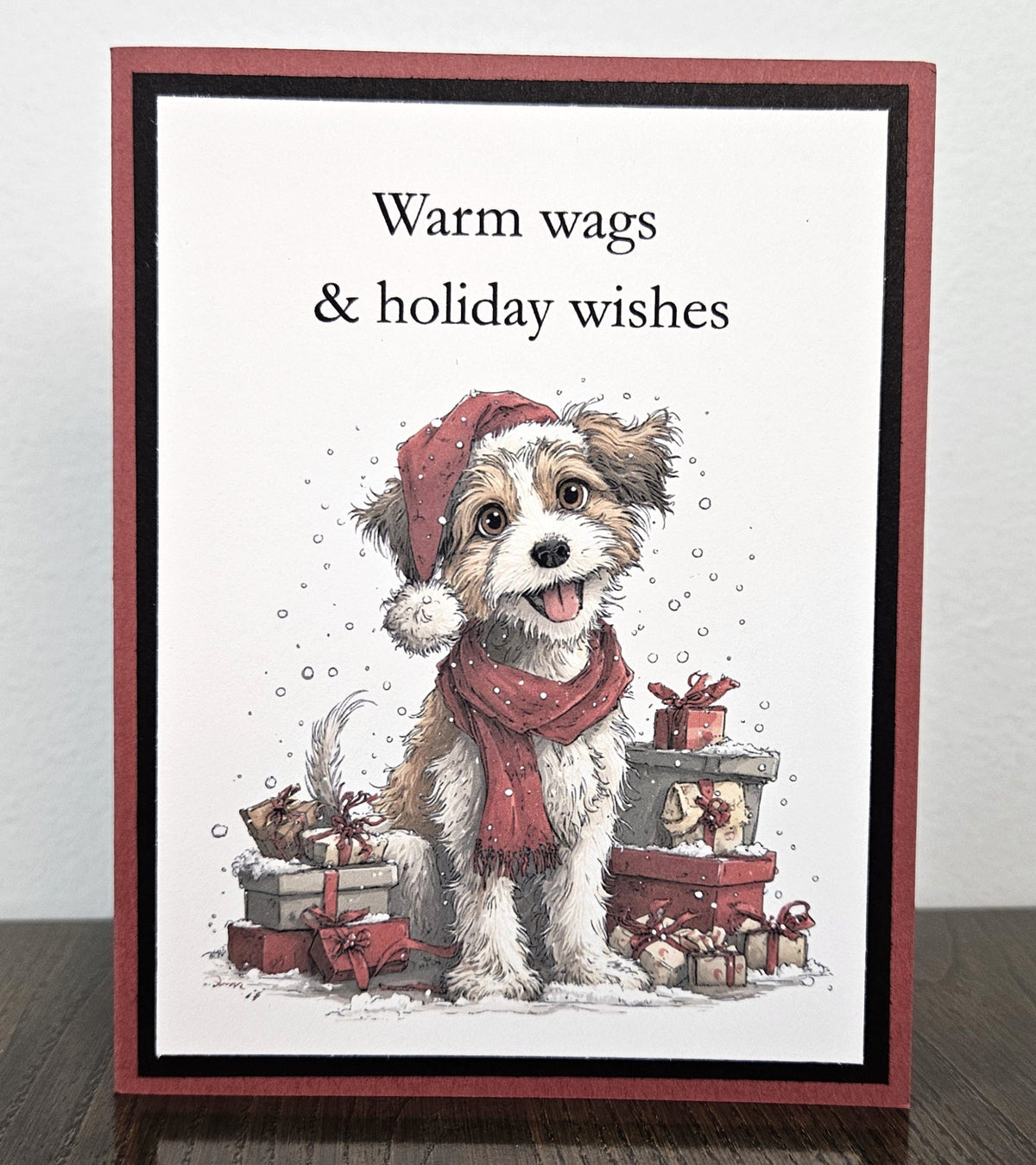 Handmade muted red and black Christmas card featuring a dog in a Santa hat and scarf among gifts, with Warm wags and holiday wishes wording.