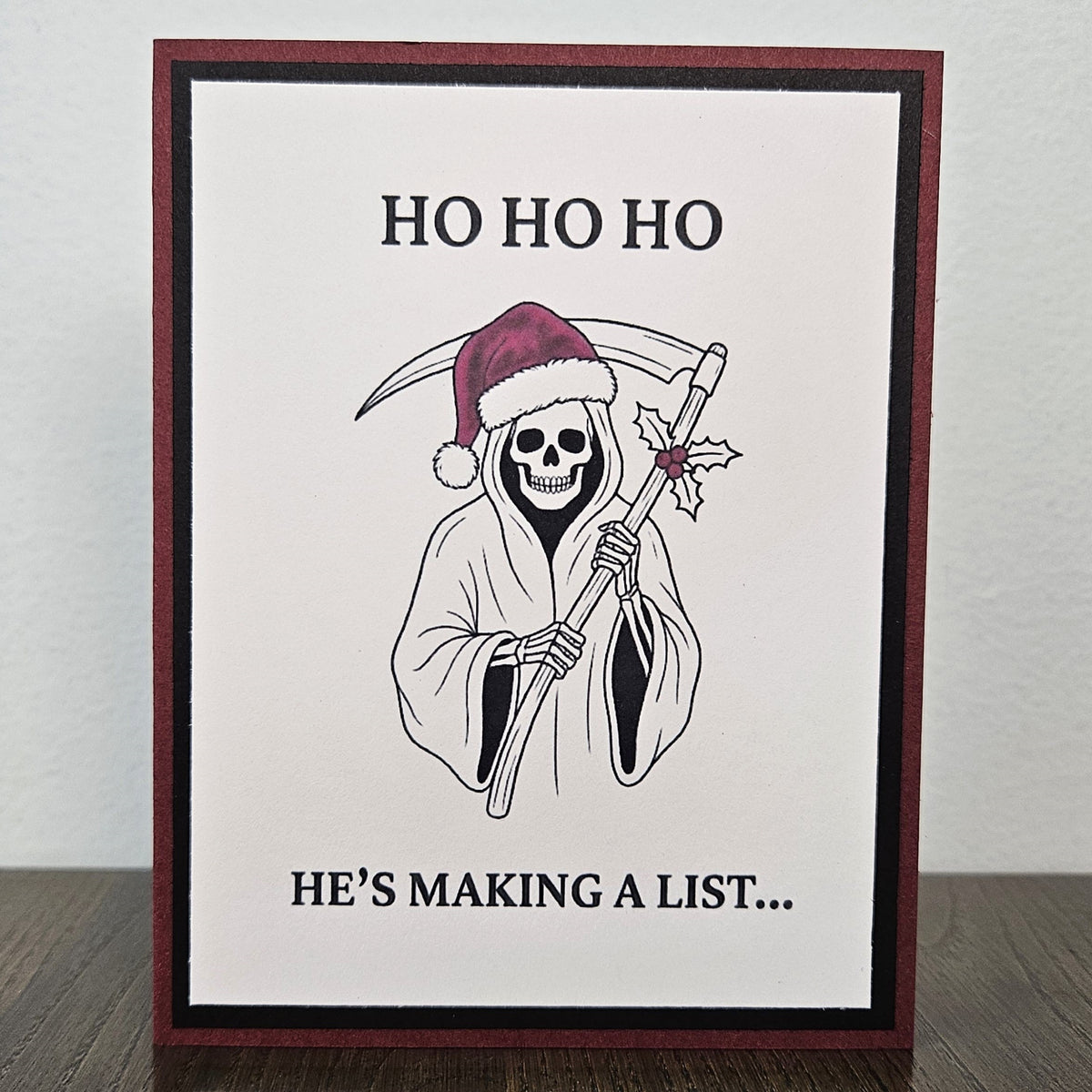 Handmade burgundy and black Christmas card featuring a Santa-hat Grim Reaper with a holly-decorated scythe and Ho Ho Ho, He's making a list wording.