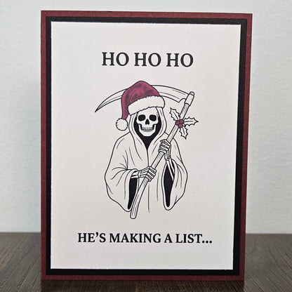 Handmade burgundy and black Christmas card featuring a Santa-hat Grim Reaper with a holly-decorated scythe and Ho Ho Ho, He's making a list wording.