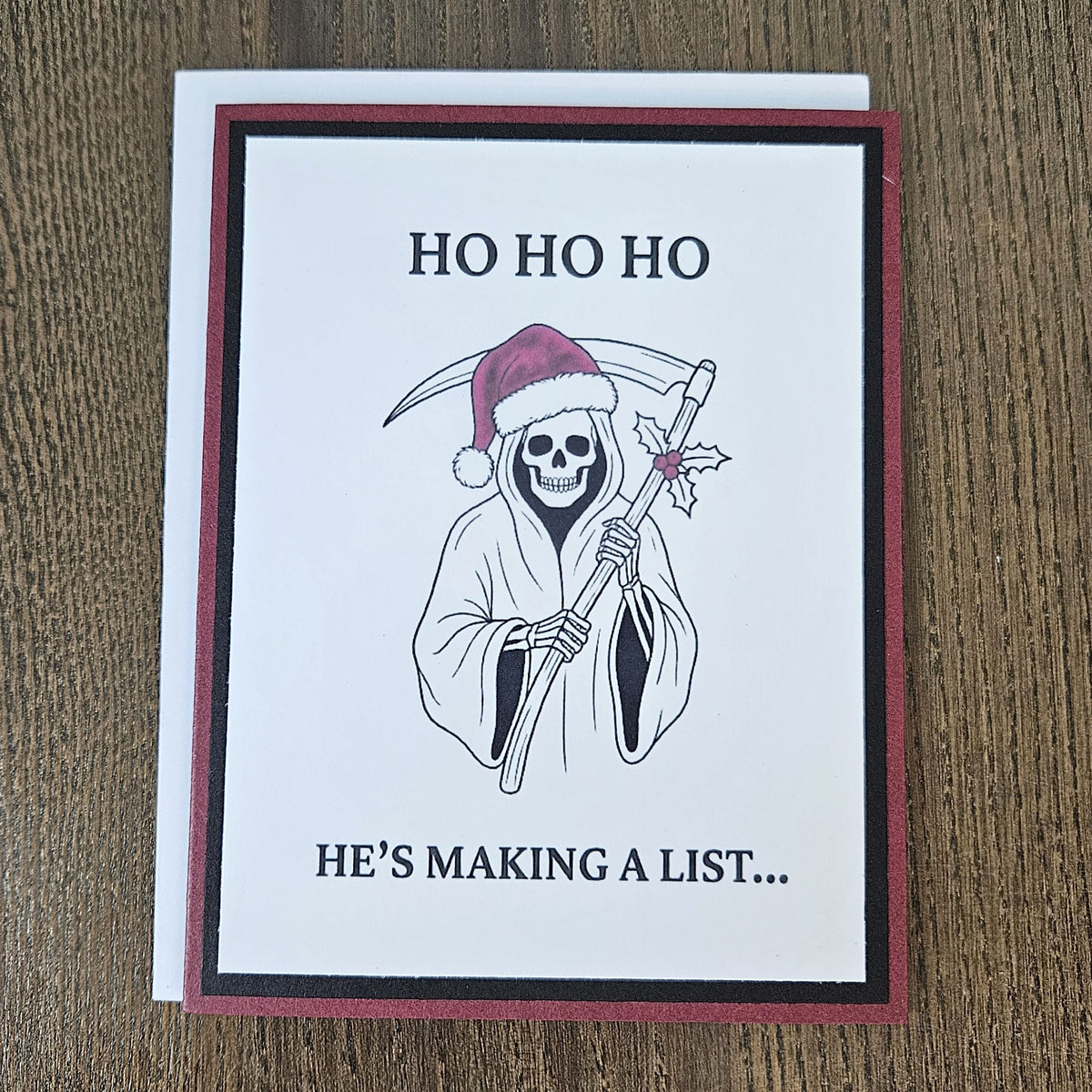 Burgundy and black Grim Reaper Santa Christmas card laid on a wooden surface with its included white envelope.