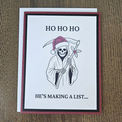 Burgundy and black Grim Reaper Santa Christmas card laid on a wooden surface with its included white envelope.