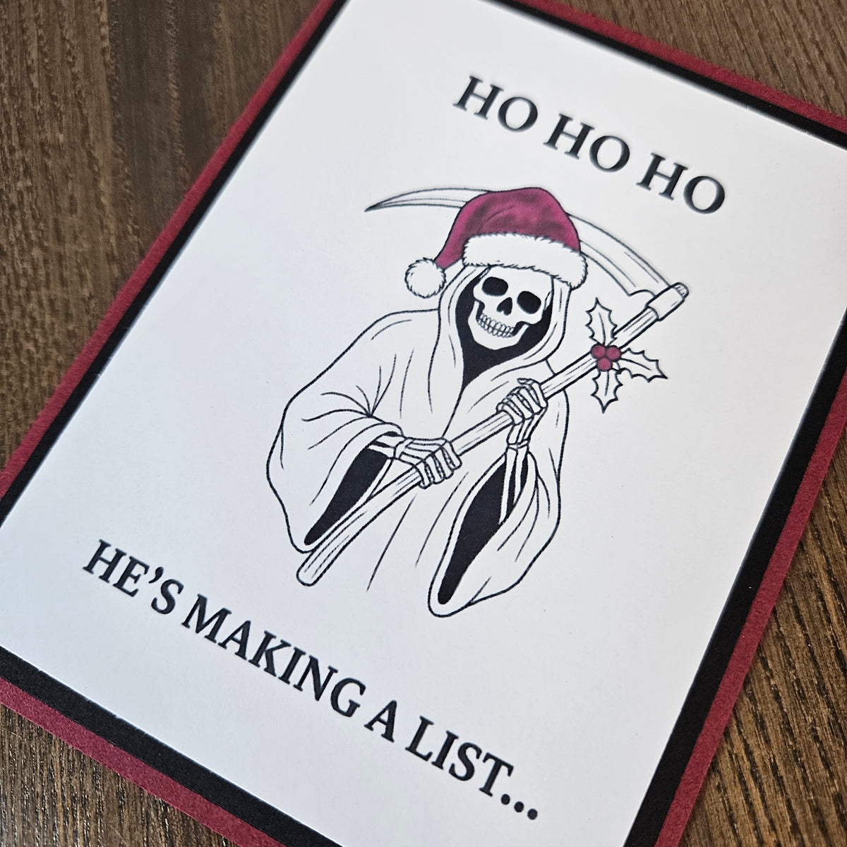Angled close-up of the printed Santa-hat Grim Reaper, holly-decorated scythe, Christmas wording, and burgundy and black cardstock borders.