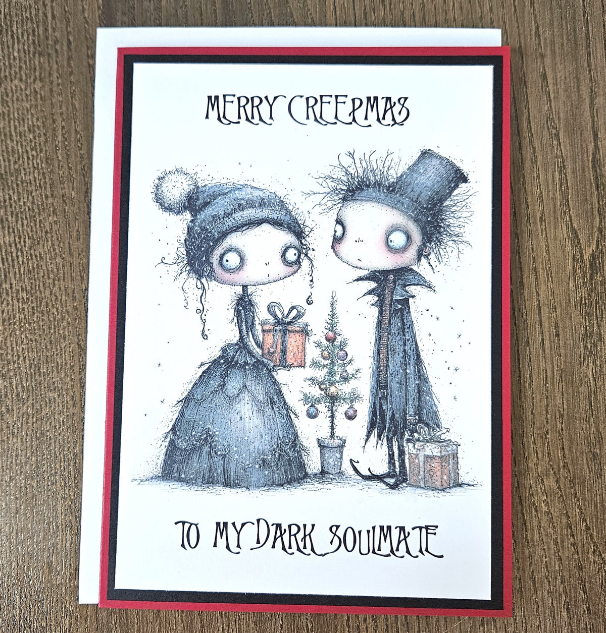 A handmade greeting card featuring a gothic-style illustration of two characters, one in a dark dress with a pom-pom hat and one in a dark coat and top hat, standing beside a small decorated tree and gifts. The card reads 'MERRY CREEPMAS' at the top and 'TO MY DARK SOULMATE' at the bottom.